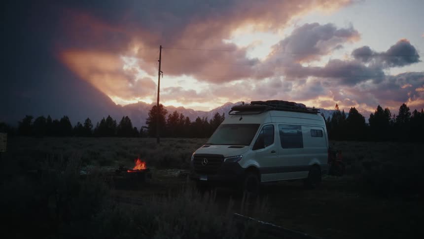 A converted camper van is parked by a warm campfire in a remote mountain meadow at dusk under a dramatic, cloudy sky