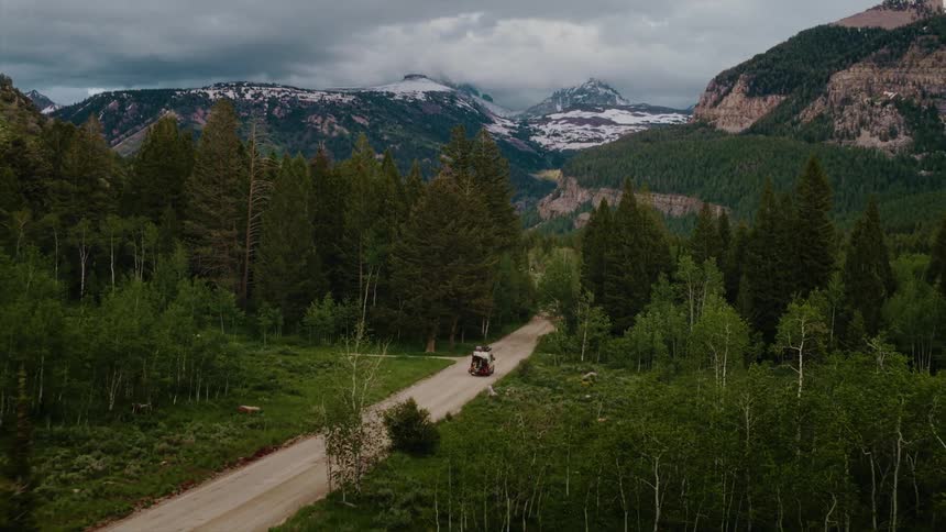 A classic car is parked on a dirt road in a lush mountain forest with snow-capped peaks in the distance