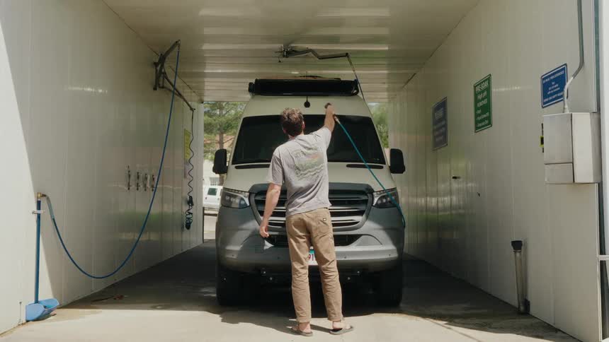 A man in sandals washes the front of his camper van with a high-pressure hose at a self-service car wash
