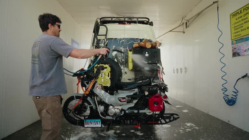 A man in slow motion uses a pressure washer to clean his adventure motorcycle and camper van at a self-service car wash