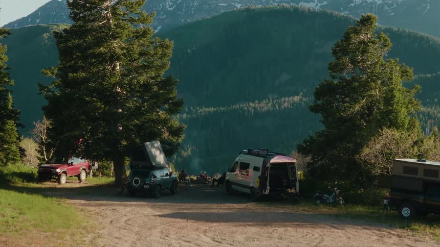 A scenic aerial shot pushes in on a group of friends gathered around their overlanding campsite in a beautiful mountain landscape