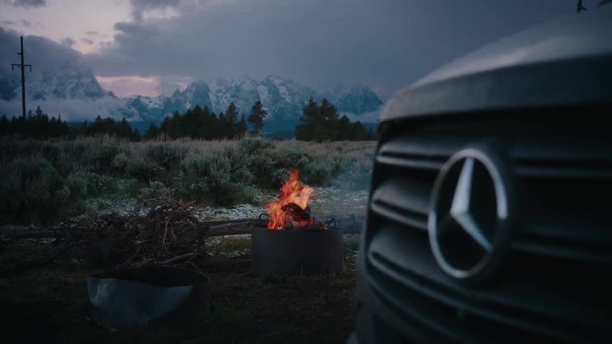 A cozy campfire crackles at a campsite next to a Mercedes-Benz van, with a majestic snowy mountain view under a dramatic twilight sky