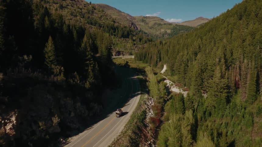 An aerial view captures a car driving along a winding mountain road nestled in a vast, lush green forest