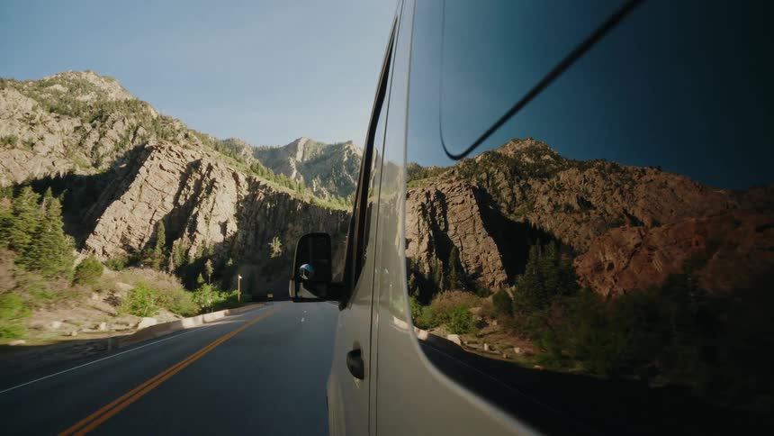 A van drives along a scenic mountain road, the rocky landscape reflected in its glossy side panels on a sunny day