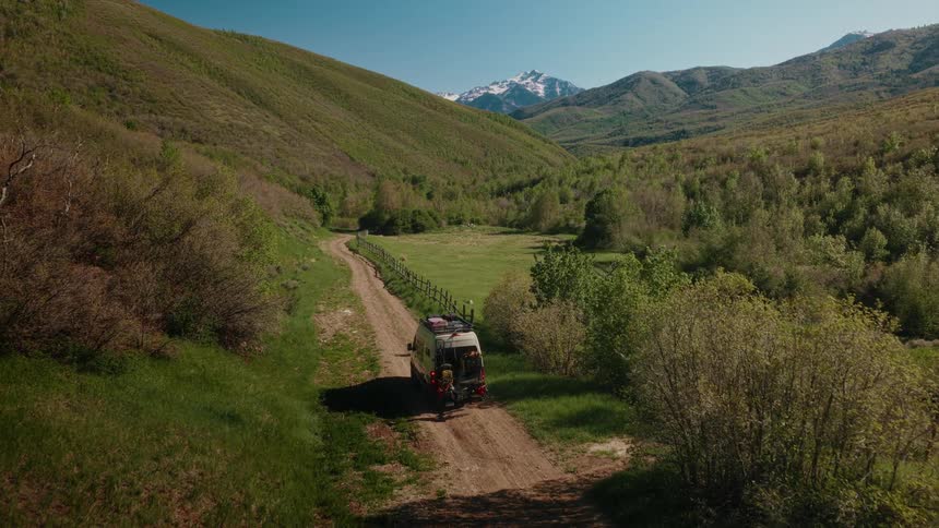 Aerial view of a camper van parked on a remote dirt road in a lush green valley with snow-capped mountains in the background