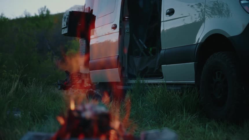 The warm glow of a campfire reflects off a nearby camper van during a peaceful evening outdoors