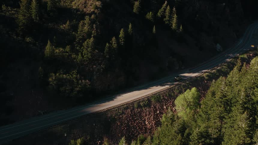 High angle aerial view of a car driving on a winding road through a lush green mountain forest during a beautiful sunset