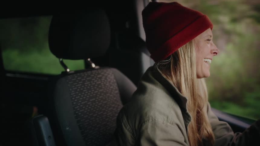A joyful woman wearing a red beanie smiles as she drives a van on a scenic road through the forest