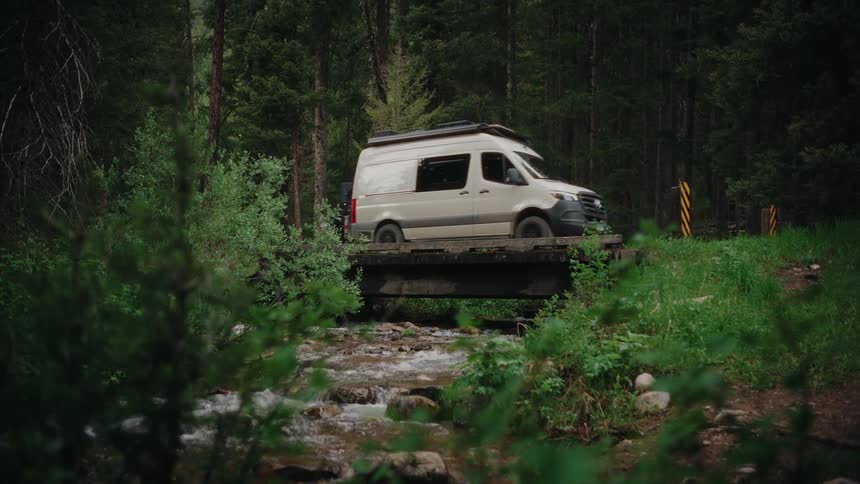 A white camper van slowly crosses a rustic wooden bridge over a flowing creek in a lush green forest