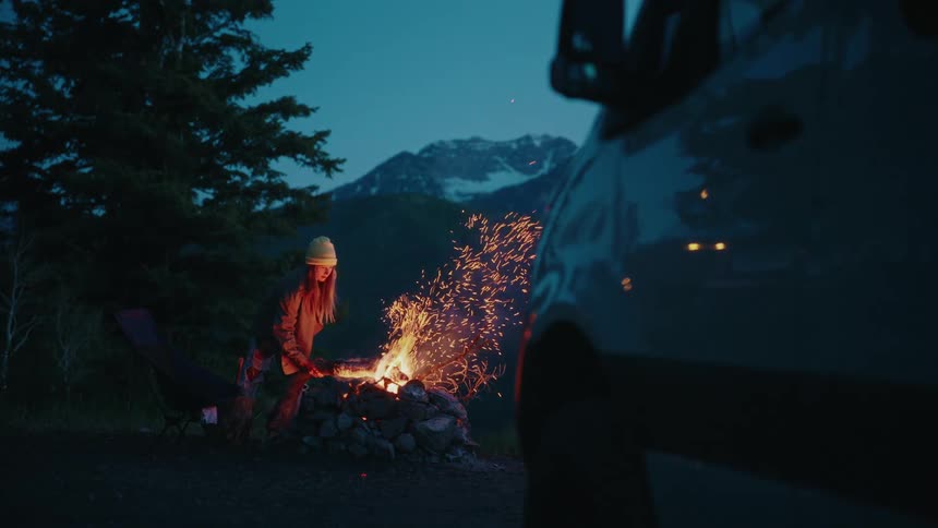 A woman adds a log to a campfire in slow motion, sending sparks flying into the night sky at a mountain campsite