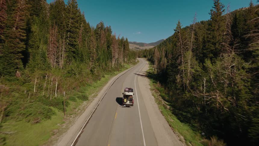 An aerial view follows a classic camper van packed with gear as it drives down a scenic mountain highway surrounded by a lush green forest