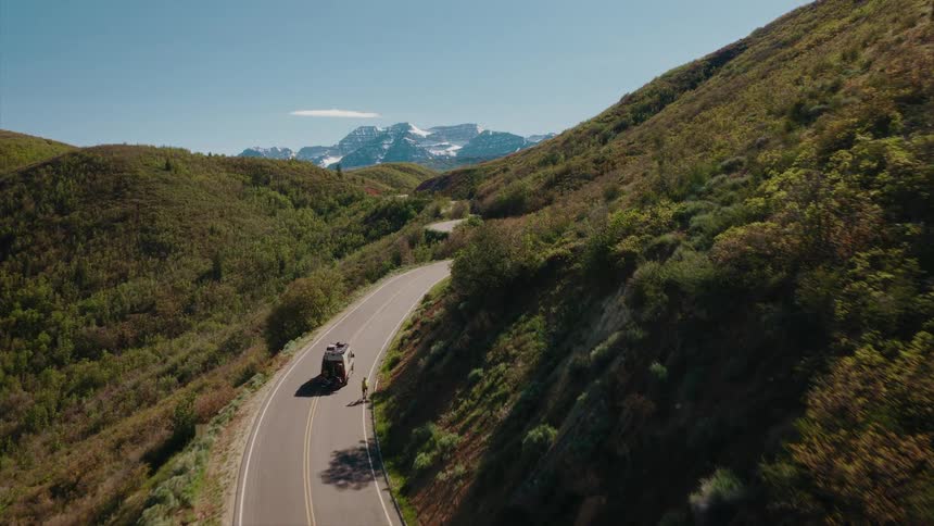 An aerial shot follows a camper van driving along a winding road through lush green hills towards a snow-capped mountain range