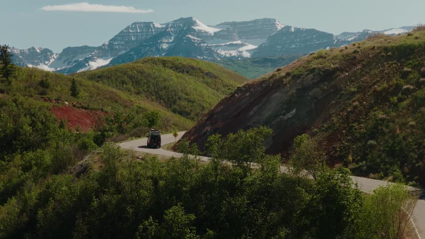 An RV drives on a scenic road trip through green hills towards a snow-capped mountain range on a sunny day