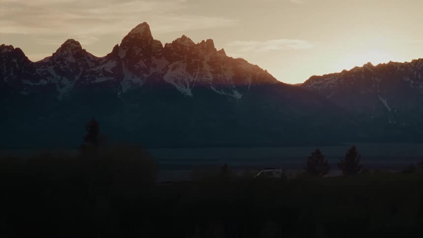 A camper van drives through a silhouetted valley against the majestic, snow-capped Grand Teton mountain range at sunrise
