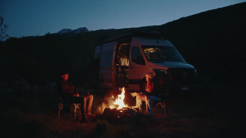 A couple enjoys a cozy campfire at night next to their modern camper van in the mountains