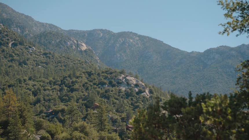 A dramatic fast zoom-in reveals the rugged, tree-covered landscape of a mountain range on a sunny day