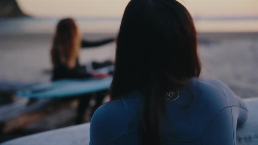 Two female surfers in wetsuits sit on the beach with their boards at sunset, one pointing towards the ocean