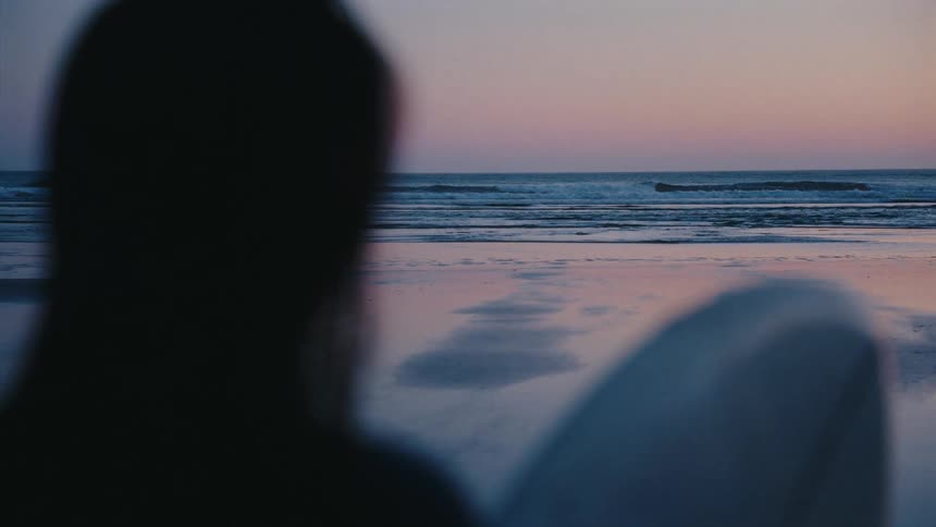 From behind, a silhouetted surfer sits with their board and watches the gentle ocean waves at sunset