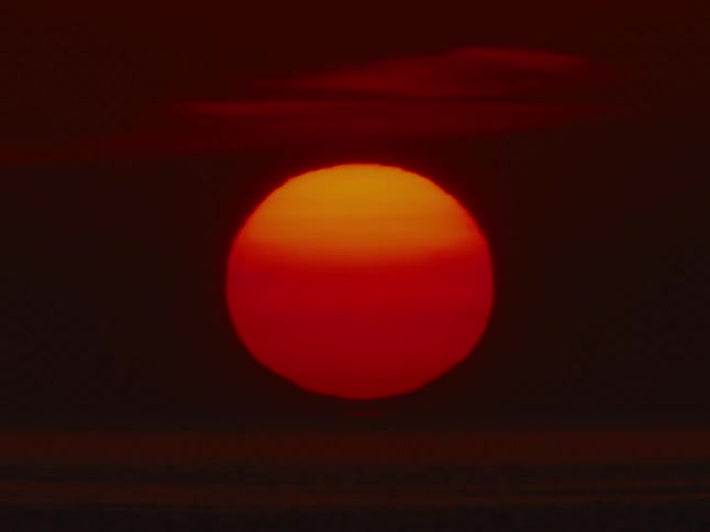 A dramatic close-up shot of a large, fiery red sun sinking below the ocean horizon