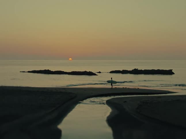 A tranquil coastal scene shows a lone surfer holding their board on a sandbar as the sun sets over the calm sea