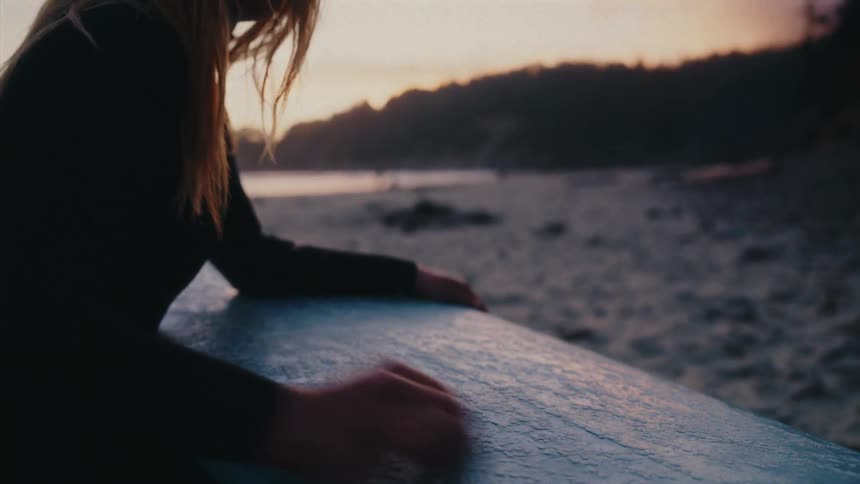 A female surfer waxes her surfboard on a sandy beach, preparing for a sunset session