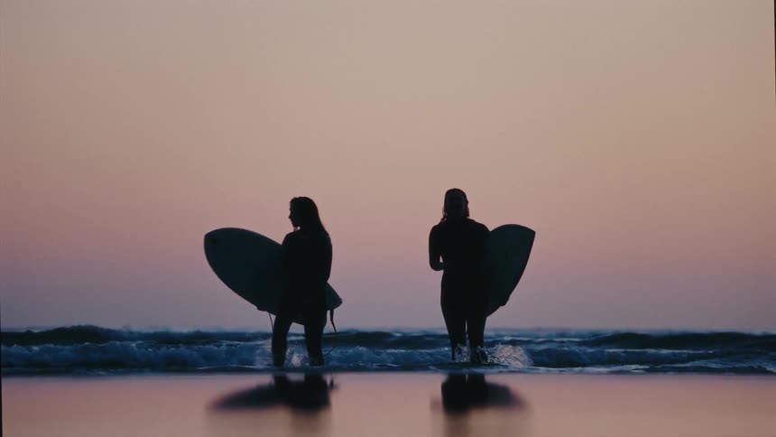 Two silhouetted women in wetsuits carry their surfboards as they walk into the ocean during a beautiful sunset