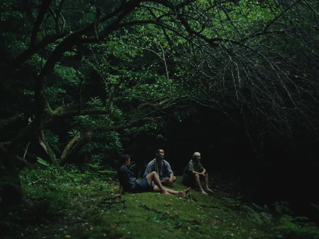Three men sit together barefoot on a lush, mossy hill while taking a break in a dark and serene forest