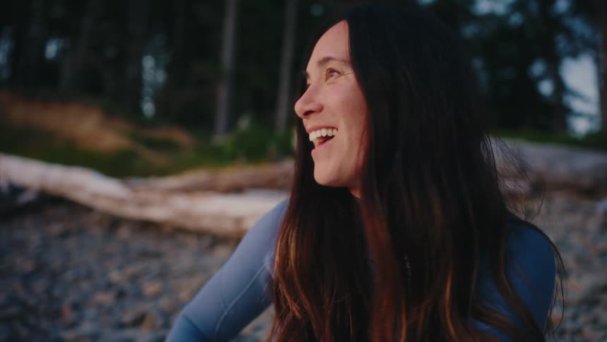 A beautiful woman in a wetsuit smiles and laughs with joy while relaxing on a rocky beach at sunset