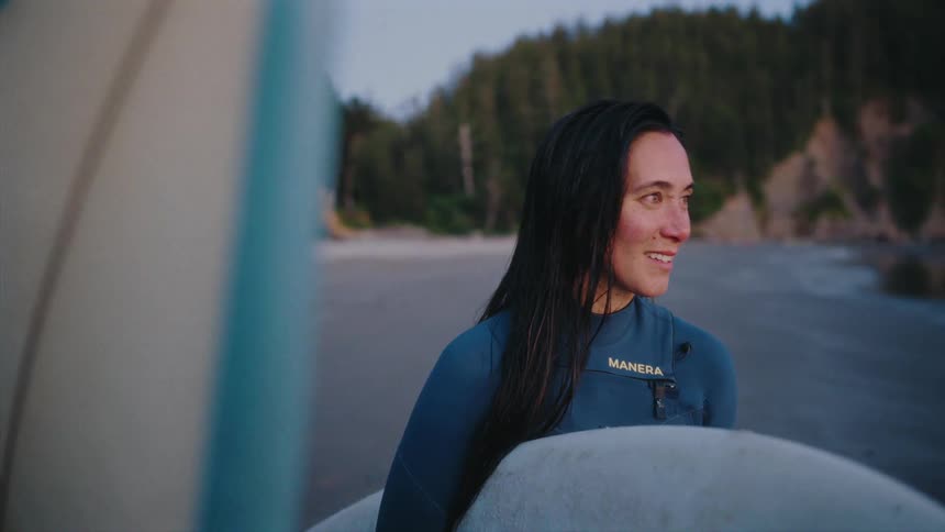 A content woman in a wetsuit stands on a remote beach with her surfboard at dusk, smiling peacefully as she looks out towards the waves
