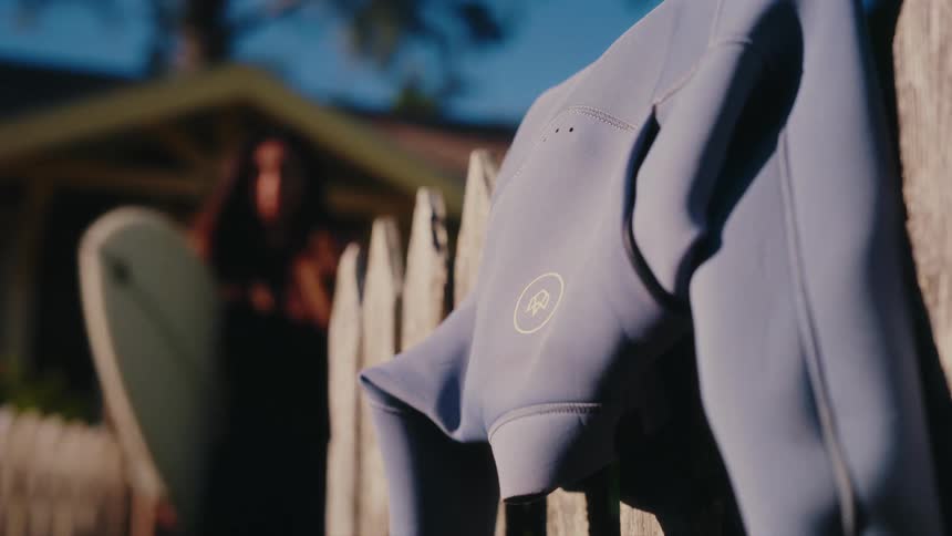 A wetsuit hangs on a wooden picket fence in the sun before a surfer grabs it and their board to head out