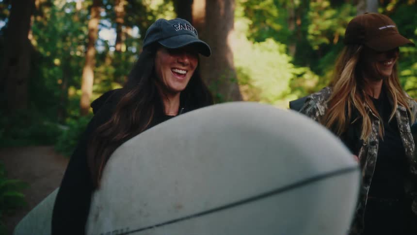 Happy surfer girls walk and laugh with their surfboards through a beautiful green forest path in slow motion