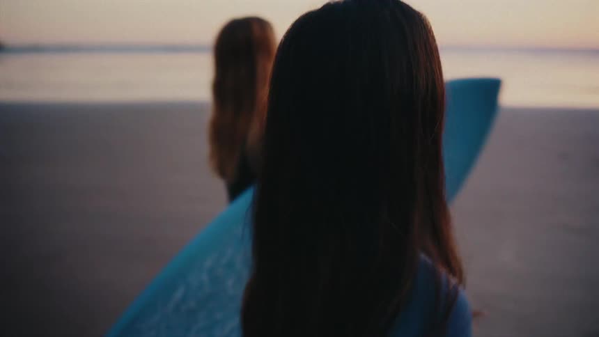 Two female surfers in wetsuits walk along a sandy beach with their surfboards during a beautiful sunset, heading towards the ocean