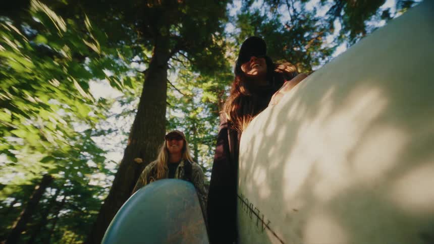 A low-angle slow-motion shot captures two smiling friends as they carry their surfboards through a lush, sunlit forest