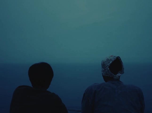 Two silhouetted people sit together looking out at a calm, misty ocean at dusk