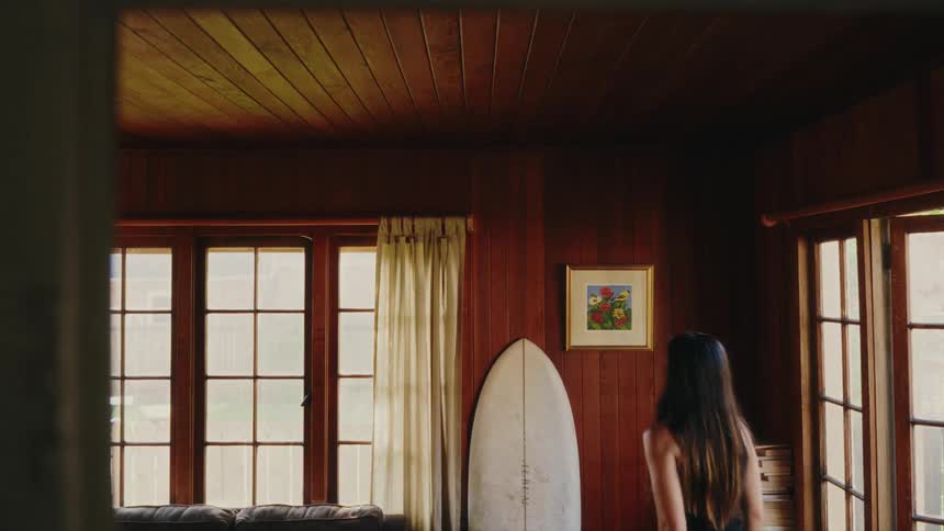 A woman with long dark hair walks into her wood-paneled living room, picks up her surfboard, and carries it out of the room