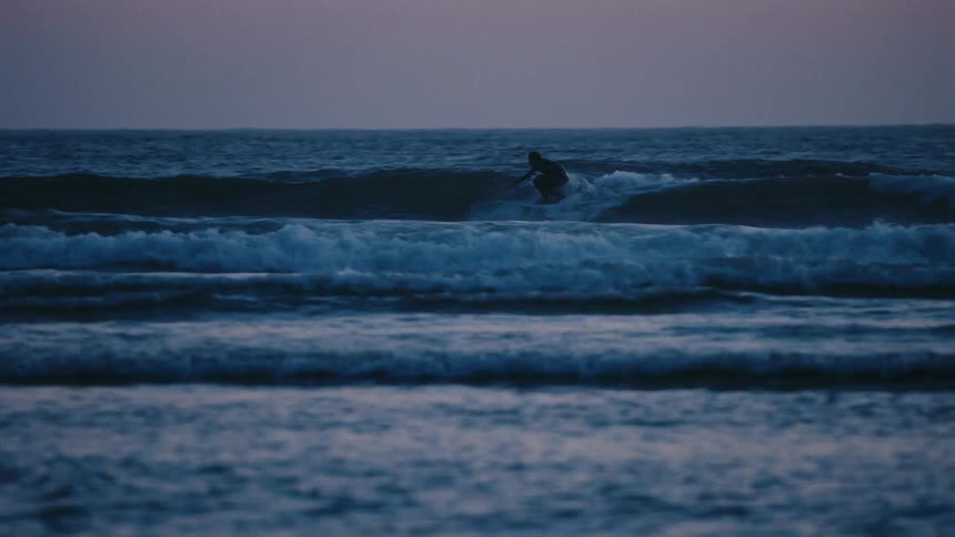 A silhouetted surfer gracefully rides a wave in slow motion across the dark blue ocean during a moody dusk