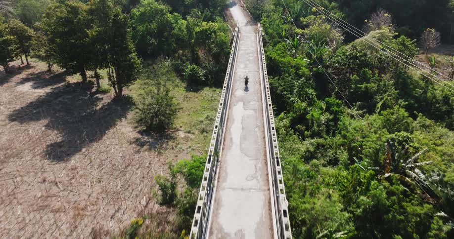 A high-angle aerial view follows a lone person riding a scooter across a long bridge over a turquoise river surrounded by a lush green landscape