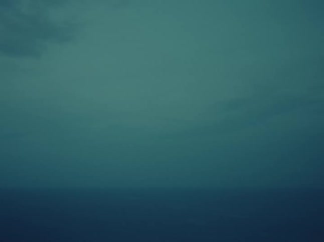 A static, monochromatic view of a dark, moody ocean meeting an overcast teal sky at the hazy horizon