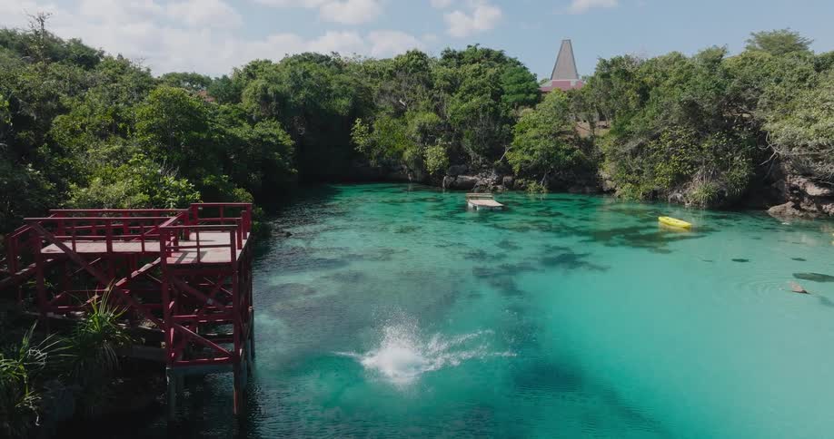 A man does a graceful backflip in slow motion from a red wooden diving platform into a crystal clear turquoise lagoon