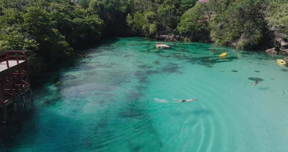 Aerial slow-motion shot of a man swimming across a stunning turquoise lagoon surrounded by a lush tropical jungle