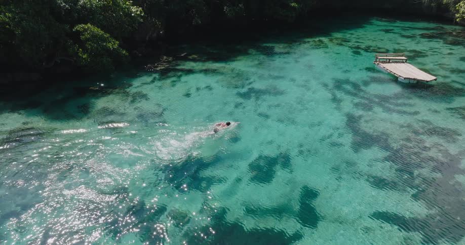 An aerial slow-motion shot captures a person swimming freestyle in the stunningly clear turquoise waters of a secluded tropical lagoon