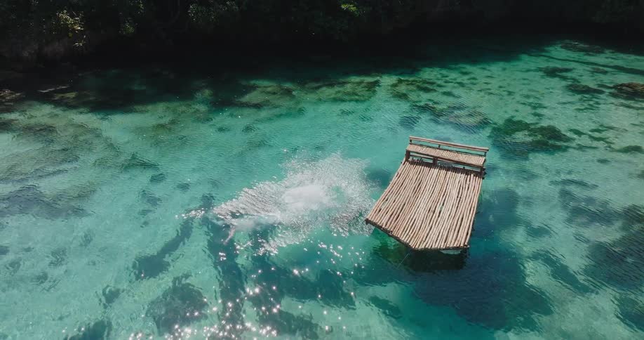 A man dives from a wooden raft into a stunningly clear turquoise tropical lagoon in slow motion, captured from an aerial perspective
