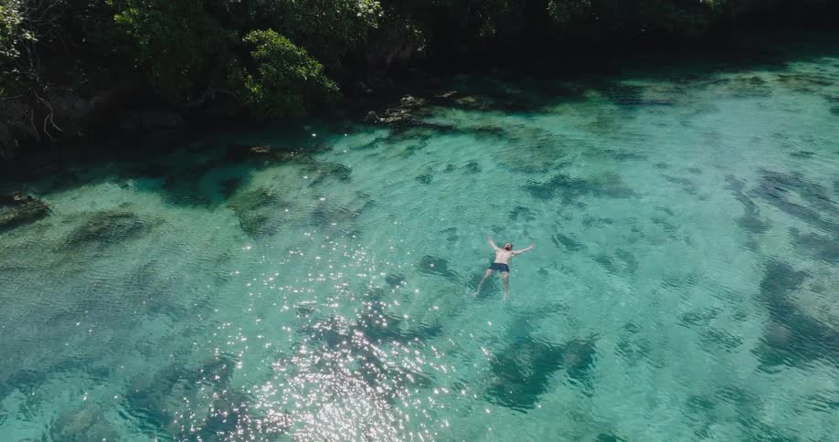 A peaceful aerial shot captures a man floating on his back and relaxing in the stunningly clear, sun-dappled turquoise ocean