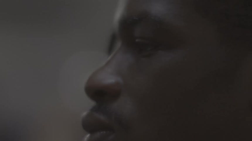 An intimate, cinematic close-up of a young Black man's face in profile, showing intense concentration in a dimly lit setting