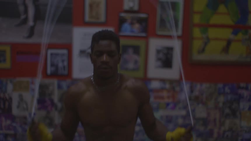 A determined shirtless boxer trains by jumping rope in slow motion in a gritty, poster-covered gym