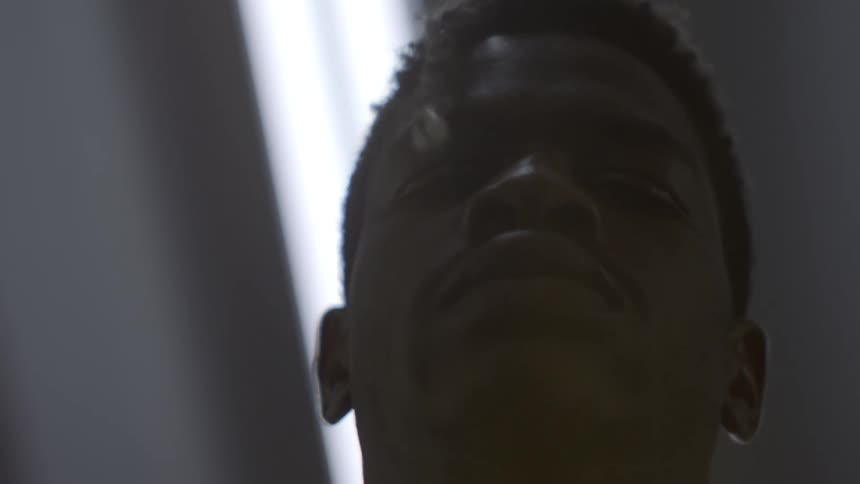An intense, low-angle close-up of a young Black man's face, silhouetted against a stark overhead light