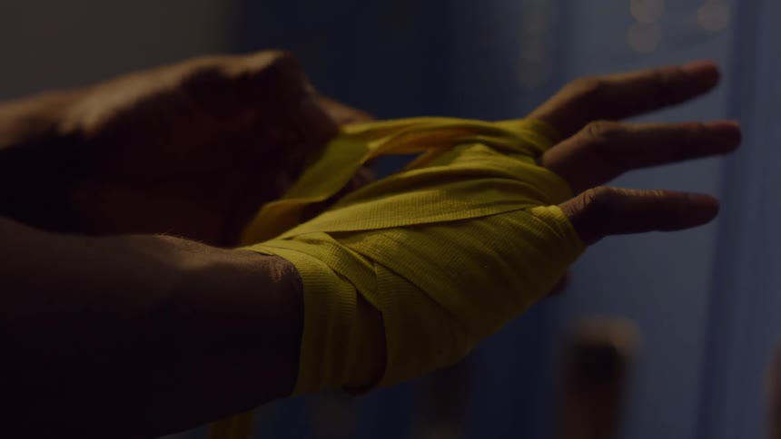 A boxer carefully wraps their hands with yellow tape in a dark locker room before training
