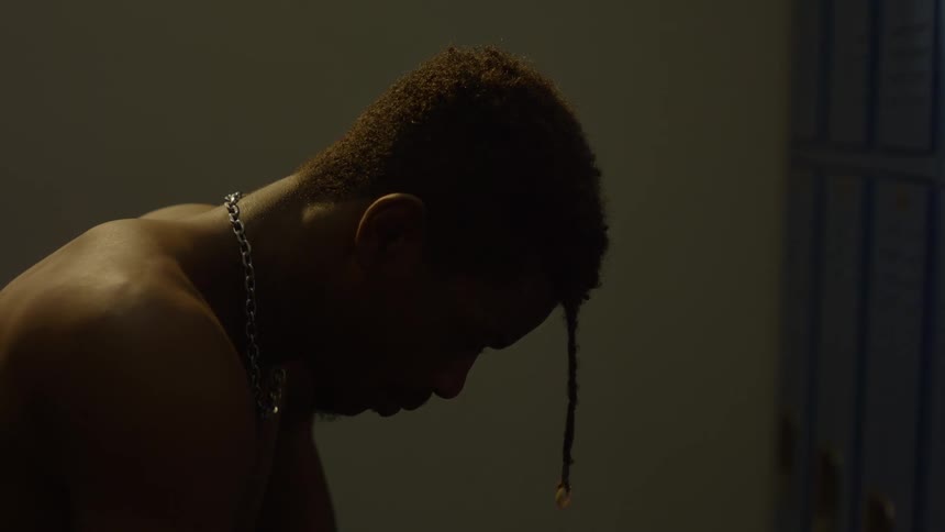 A shirtless Black athlete sits in a dark locker room and hangs his head in contemplation