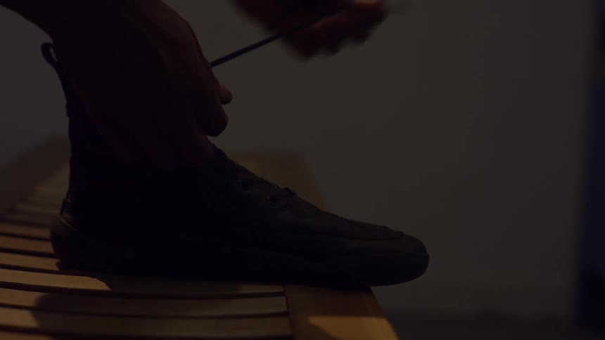 A person ties the laces on a black boot while sitting on a wooden bench in a dimly lit, atmospheric room