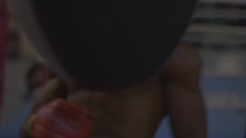 A focused, shirtless boxer practices his punches on a heavy bag in a gritty gym setting in slow motion
